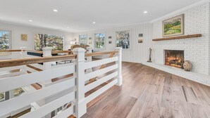 A spacious living room with a white brick fireplace, wooden flooring, and a white railing. - Waterfront Retreat with Private Dock and Fire Pit  |  Sleeps 8, Outdoor Dining, Spa-Style Primary, Chef’s Kitchen w/ Dual Dishwashers, Gas Fireplace, River Views, Bright Open Layout, Central A/C, Perfect Coastal Escape for Families, 24 Hr Hospitality (Falmouth)