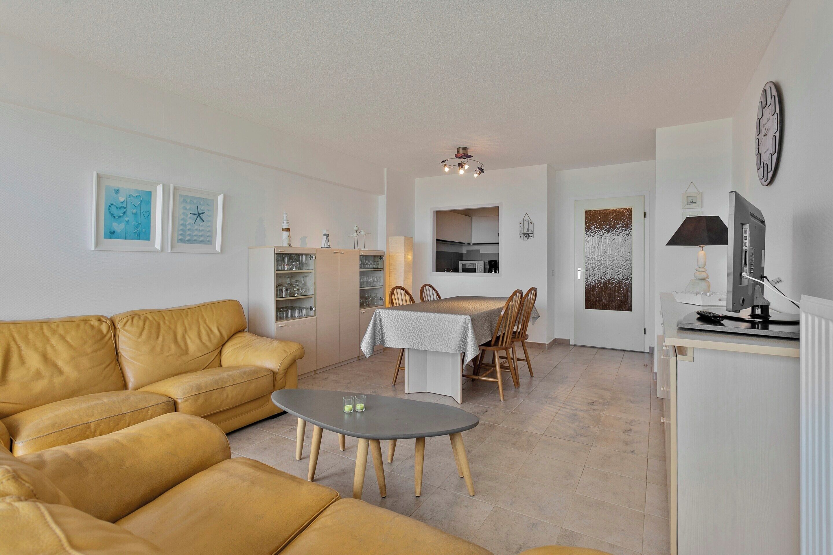 A modern living room with a sofa, a dining table, and chairs. There is a kitchen area visible in the background.