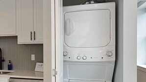 Un lavadero pequeño y blanco, con lavadora y secadora integradas, una encimera con dispensador de jabón y vistas al área de la cocina contigua.