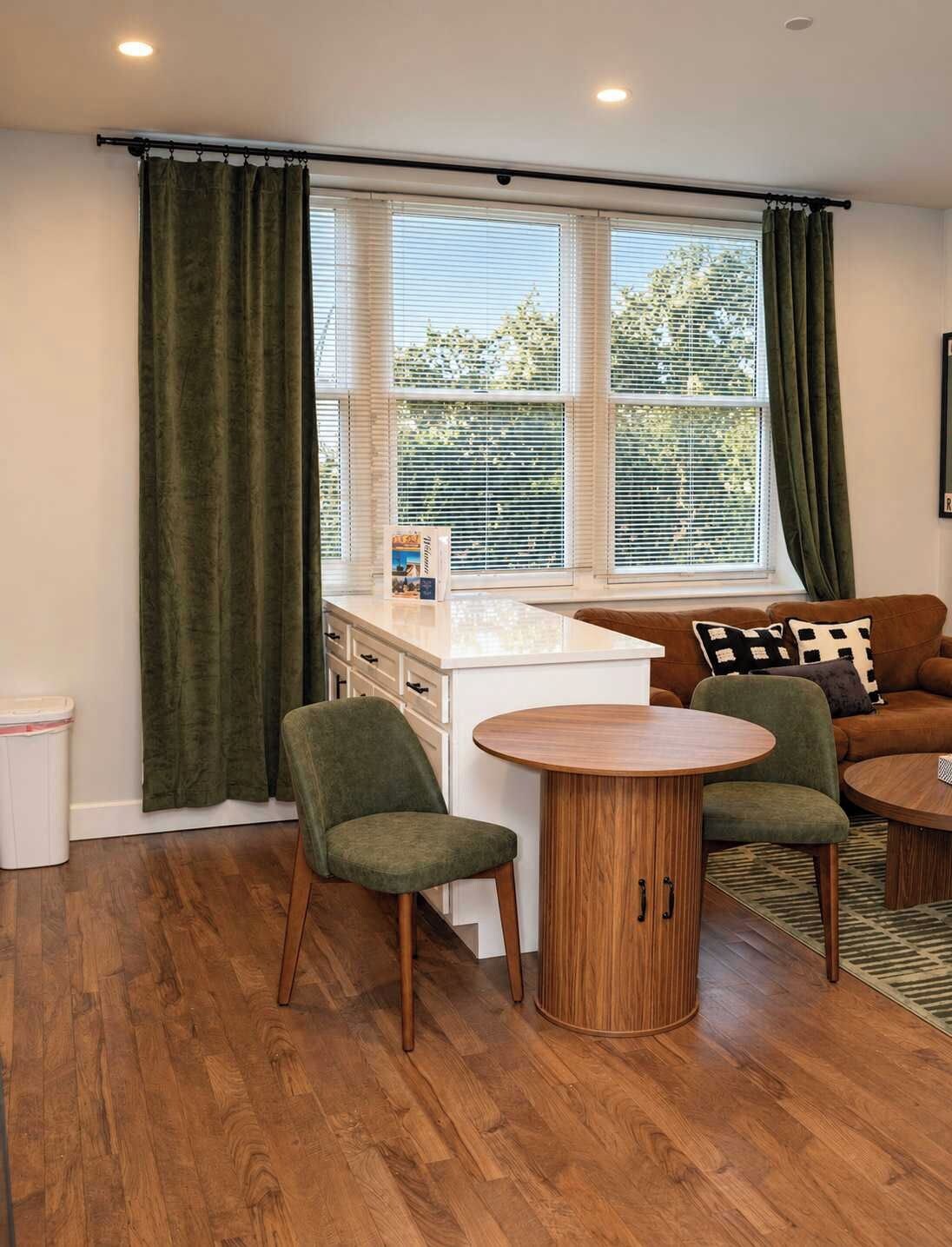 Un salon doté d’un plancher en bois, d’une petite cuisine blanche, d’une table ronde en bois, de deux fauteuils verts et d’un canapé marron agrémenté de coussins à motifs.