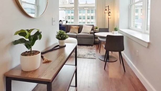Una sala de estar moderna con un sofá, una mesa de centro y una zona de comedor. Hay un espejo grande en la pared y una planta en maceta sobre una mesita auxiliar.