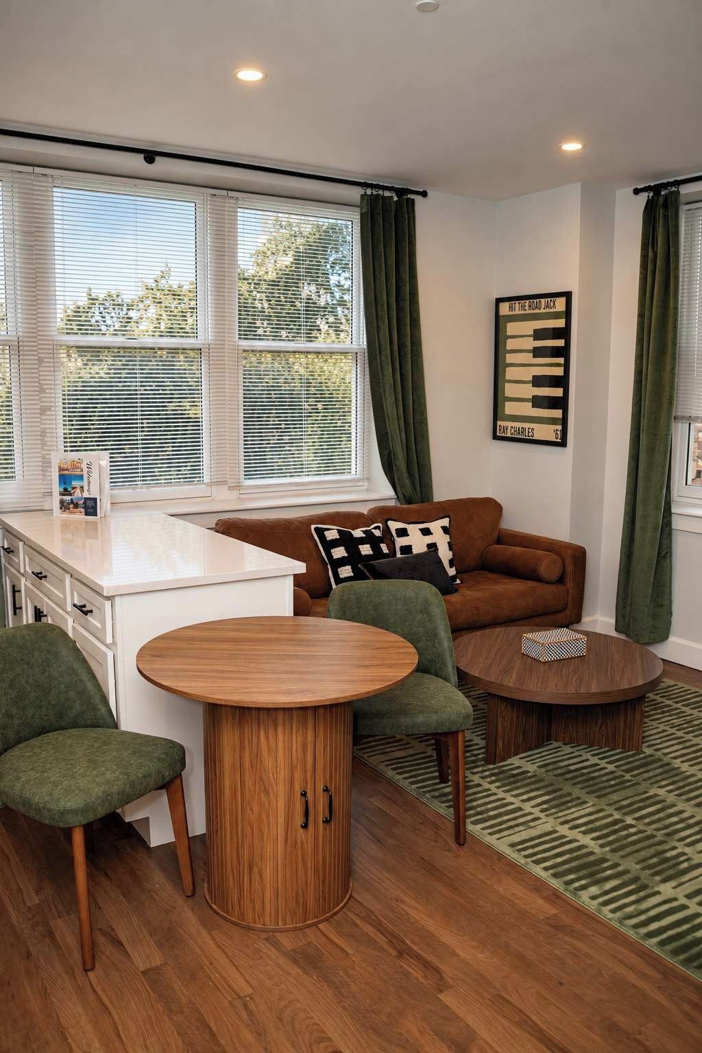 Un espacio de estar compacto con kitchenette, un sofá marrón, un sillón verde, dos mesas de madera y una alfombra estampada.