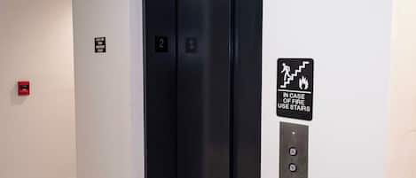 Un ascensor moderno con puerta negra, un detector de incendios y un cartel que indica el uso de escaleras en caso de emergencia.