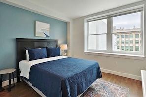 A bedroom with a large bed, a small round table, a window with a view of buildings, and a patterned rug.
