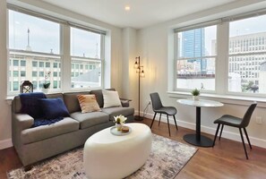 Una sala de estar moderna con un sofá gris, un reposapiés blanco y una mesa redonda con dos sillas. Amplias ventanas ofrecen vistas del paisaje urbano.