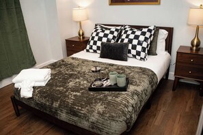 Ein ordentlich bezogenes Bett mit karierten Kissen, ein Tablett mit Tassen und einer Teekanne sowie zwei Nachttischlampen.