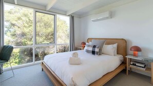 Ein modernes Schlafzimmer mit großem Fenster, einem Bett mit weißen Bettwäsche, einem grünen Sessel und einem Nachttisch mit Büchern und einer Lampe.