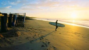 Eine Strandansicht mit einer Person, die einen Surfbrettragen, einer Treppe und einem klaren Himmel.