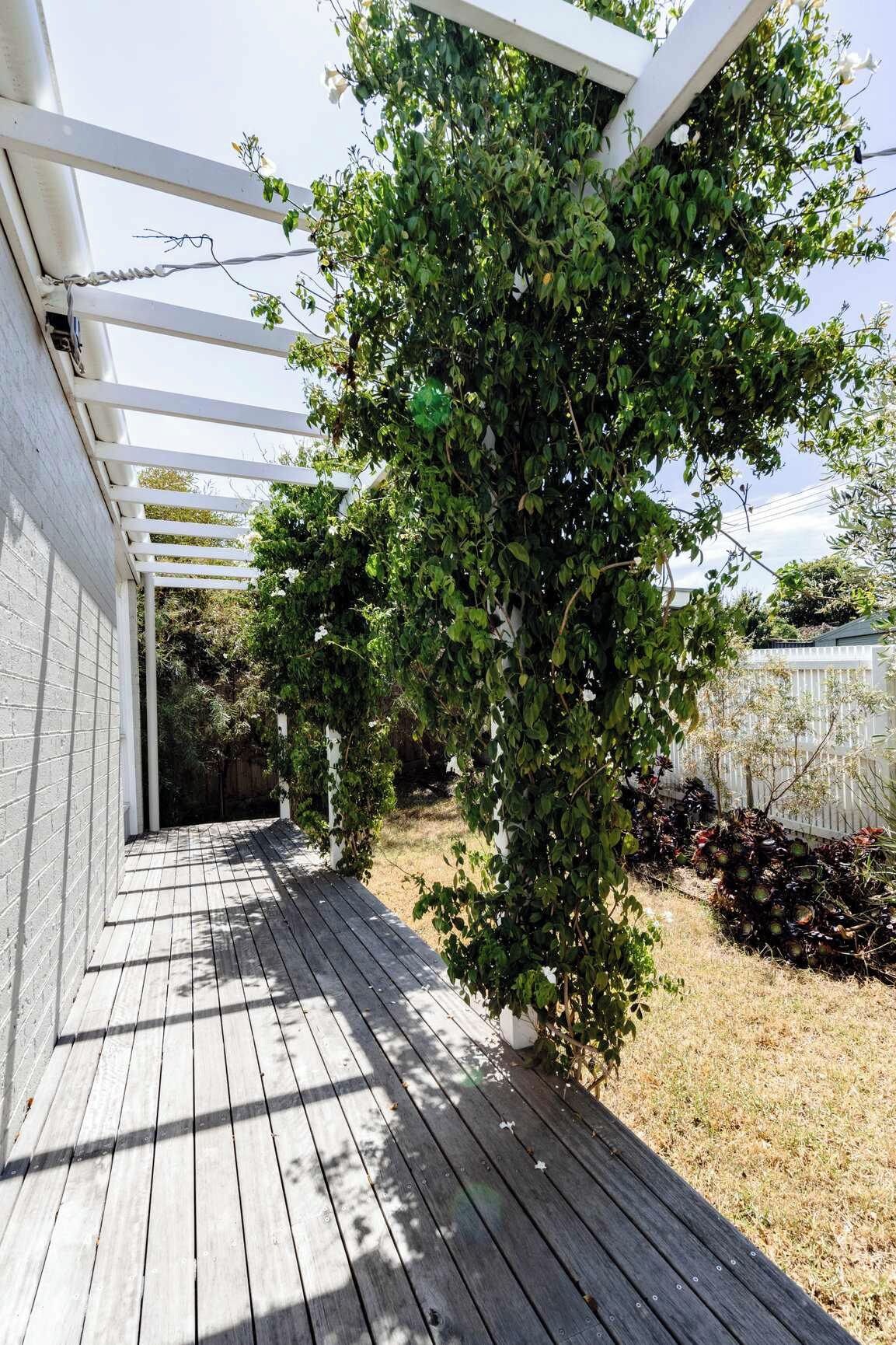 Um deck de madeira com estrutura de pérgola branca, rodeado por vegetação e um muro branco.