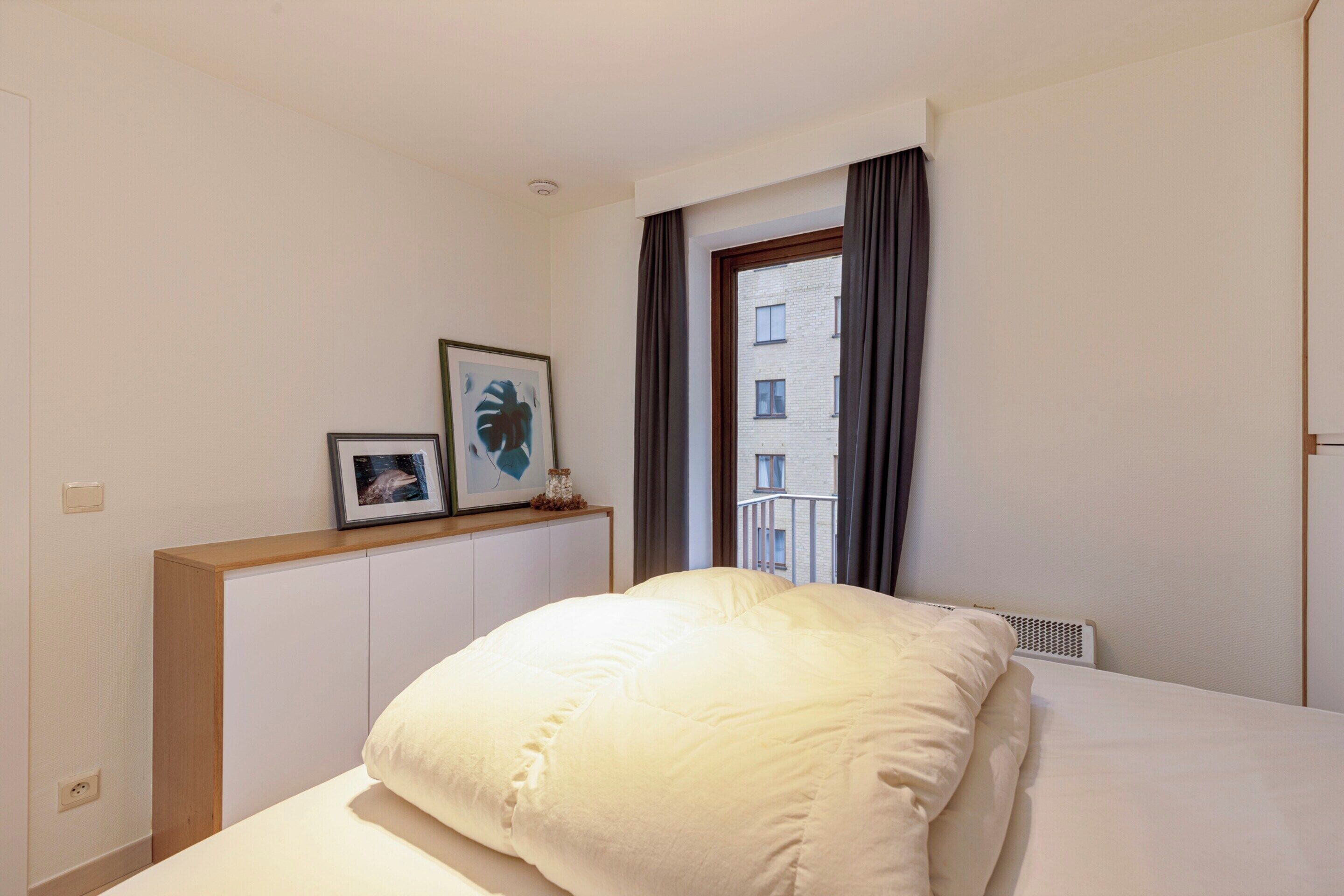 A hotel room with a bed, a window with curtains, a wall-mounted shelf with framed pictures, and a radiator.