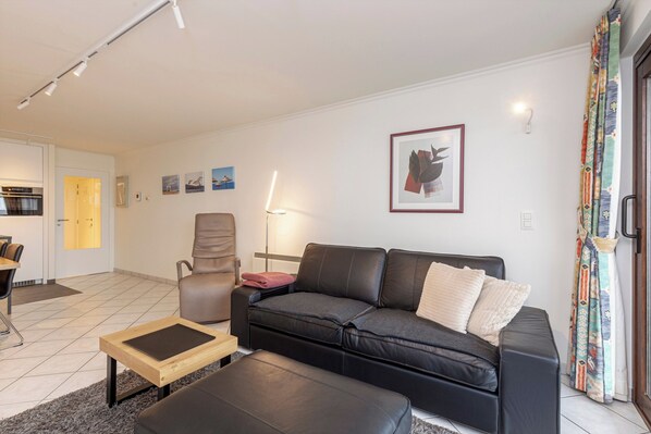 A modern living room with a black leather sofa, a wooden coffee table, and a beige armchair. There is a framed abstract painting on the wall, a lamp, and a colorful curtain. - Apartment in Blankenberge by Marina Views (Blankenberge)