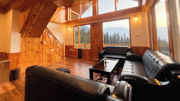 A spacious loft-style room with a wooden interior, a staircase, a seating area with a sofa and coffee table, and large windows offering mountain views. - Singing Woods Kutla (Bhuntar)