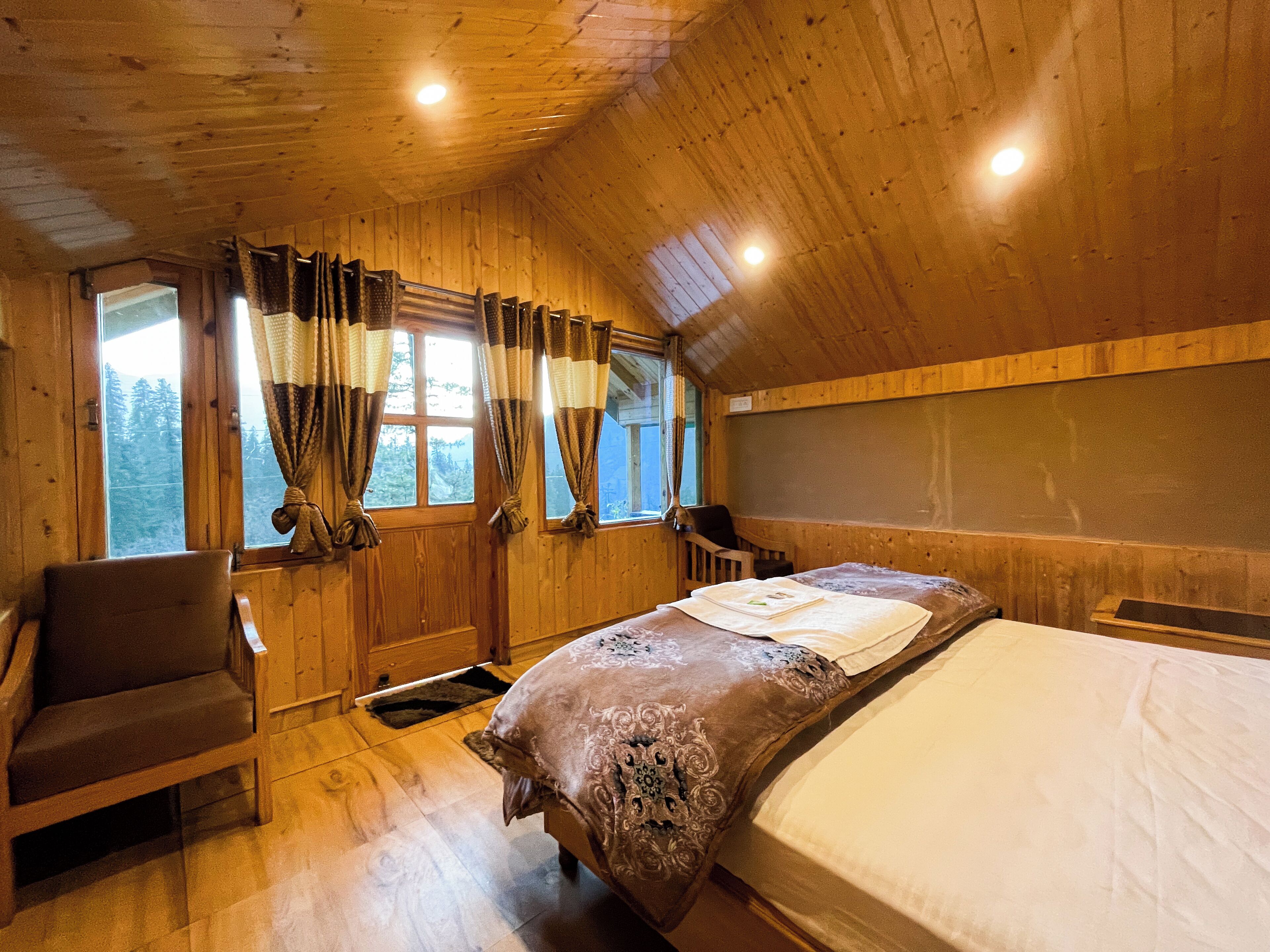 A wooden cabin room with a bed, a chair, and a window with curtains.