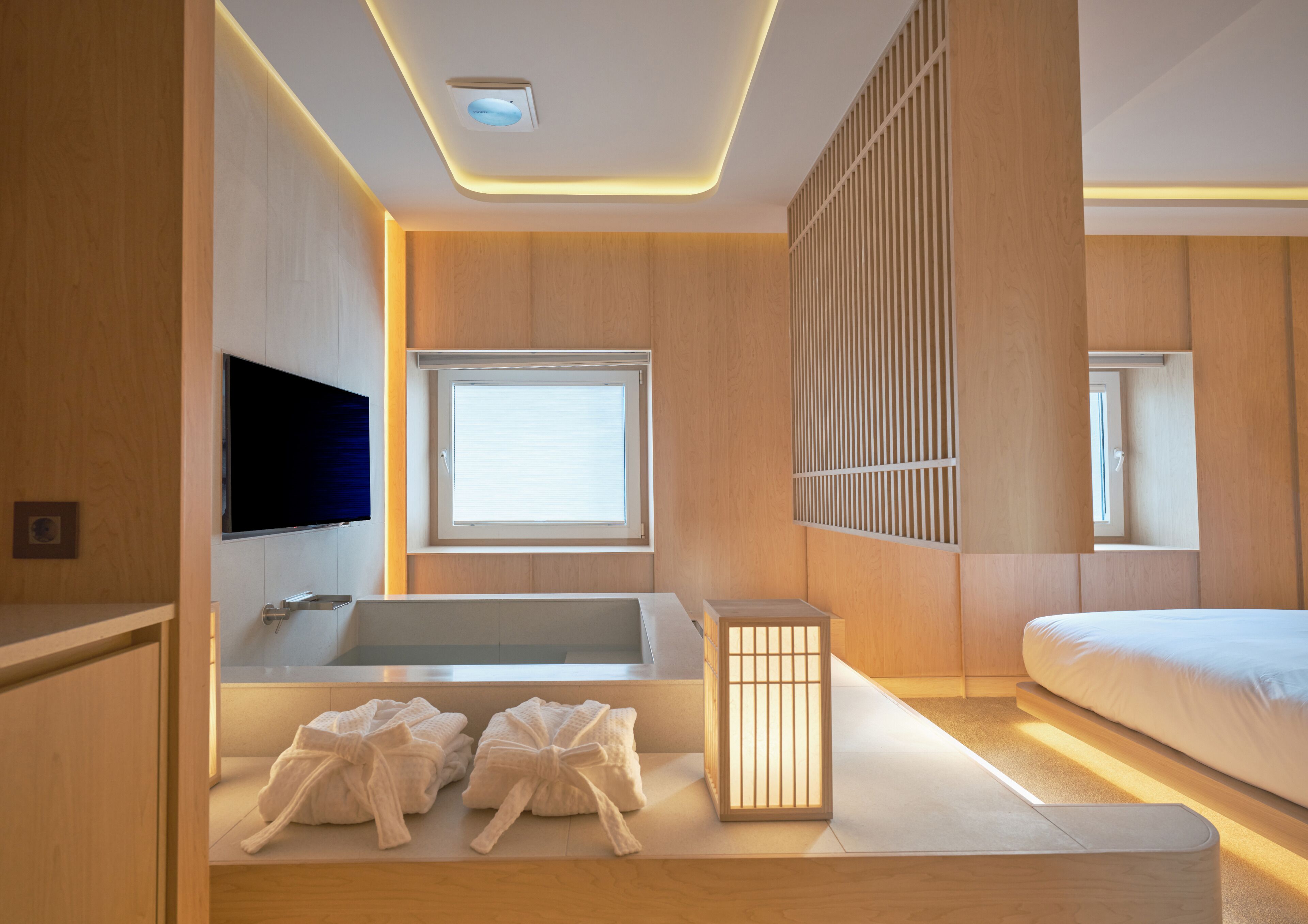 Un baño moderno con bañera, ducha e inodoro. Hay una ventana que proporciona luz natural y un televisor montado en la pared.