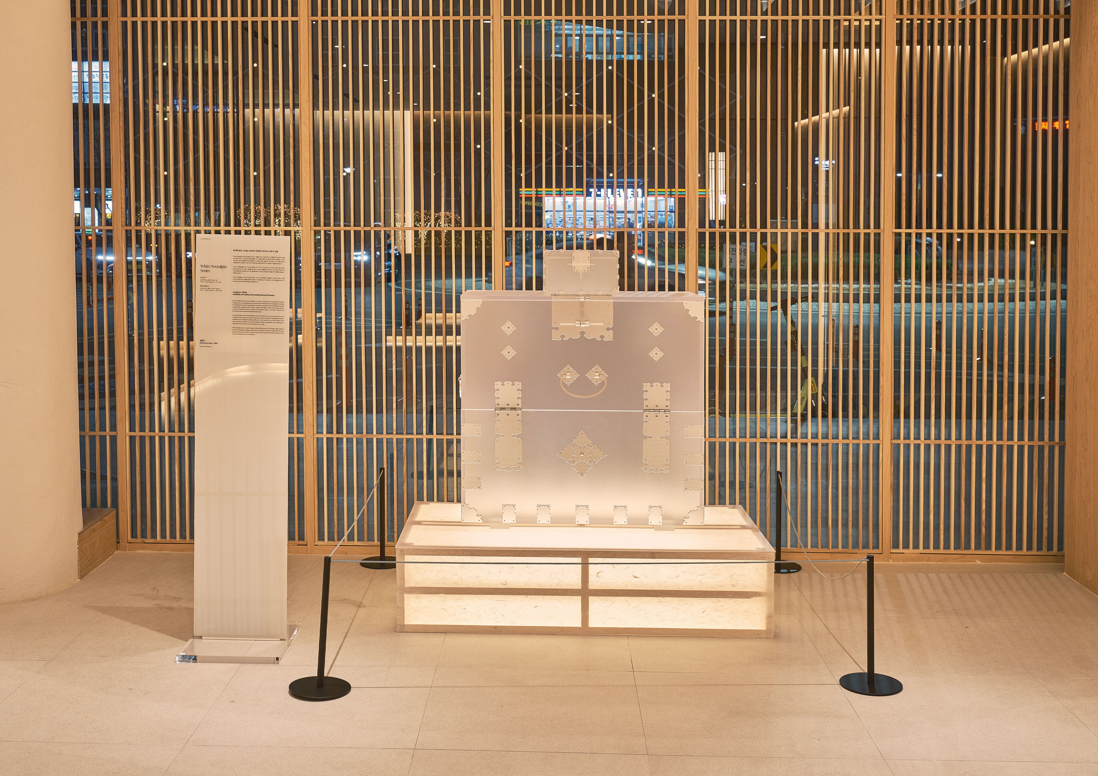 Exposição de um conjunto de chá japonês tradicional em um museu, com um edifício moderno visível através de uma janela de treliça de madeira ao fundo.