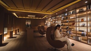 A modern library with a woman reading in a leather chair, wooden bookshelves, and a warm ceiling design. - UH Continental CenterPoint (Busan)