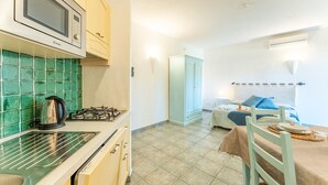Een kleine keuken met een magnetron, waterkoker en gasfornuis. Een eethoek met een tafel en stoelen is zichtbaar.