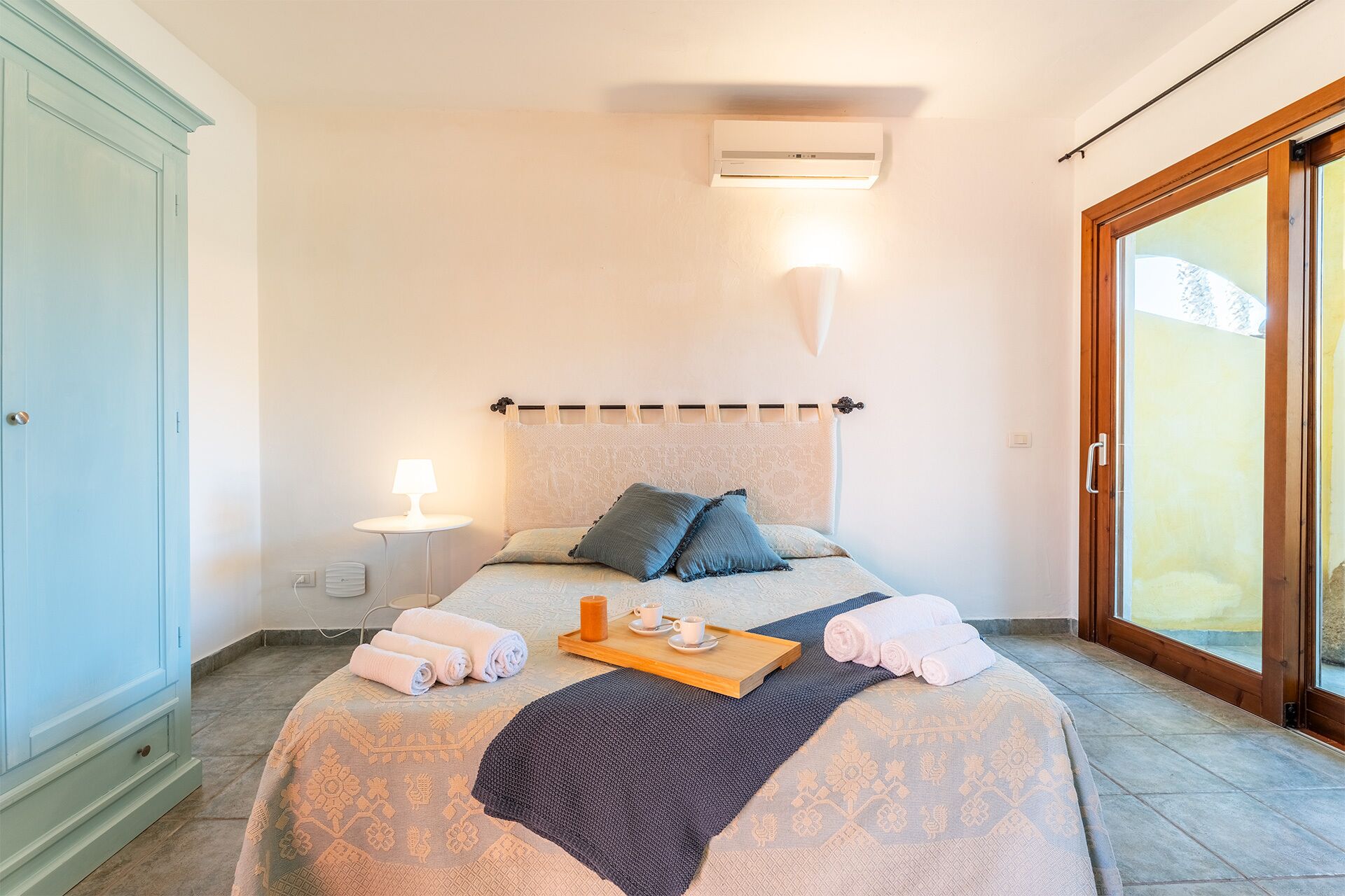 Un dormitorio con cama, mesita de noche, lámpara y una bandeja con toallas y una taza.