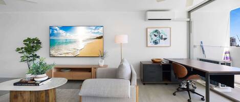 A modern living room with a beach-themed painting, a round wooden coffee table, a grey sofa, a desk with a computer, and a standing lamp.
