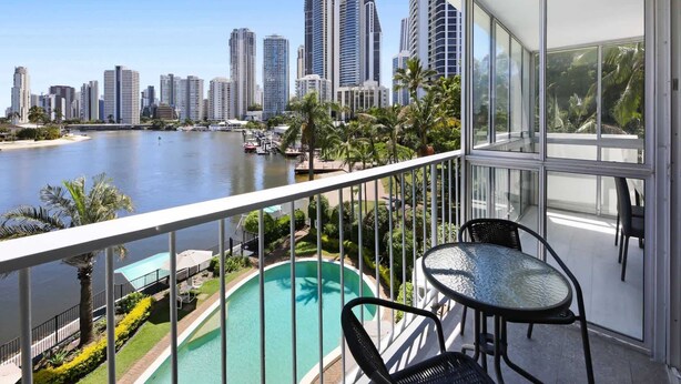 Un balcón con barandilla de vidrio con vista a una piscina y un río, con edificios altos al fondo.