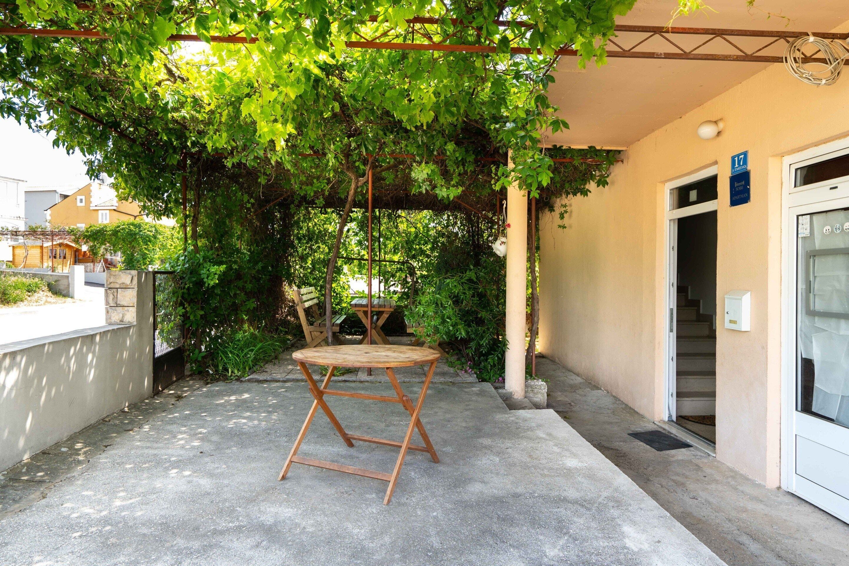 A concrete patio with a wooden table and chairs under a pergola with green vines.