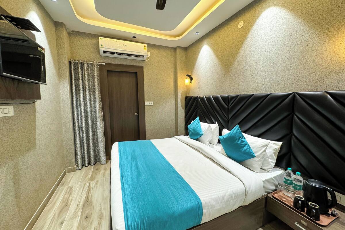 A hotel room with a bed, a TV, an air conditioner, a nightstand with a coffee maker, and a wall-mounted light.