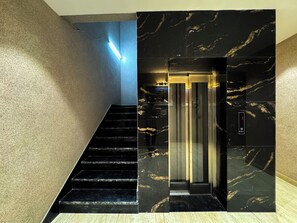 A modern elevator with a sleek black and gold marble finish, a staircase with black steps, and a textured beige wall. - Wanna stay haridwar  (Haridwar)