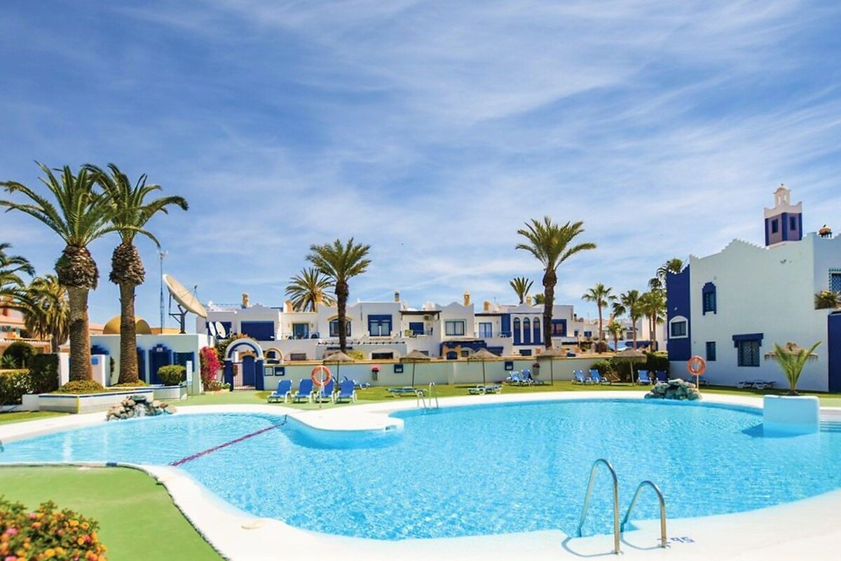 A swimming pool with lounge chairs and palm trees, surrounded by buildings with a Mediterranean architectural style.