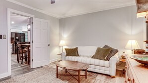 Una sala de estar con sofá, mesa de centro y lámparas. Hay una puerta que conduce a una cocina y comedor.