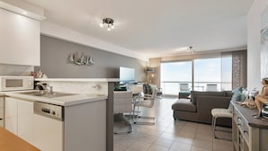 A modern kitchen with a central island, a dining area with a table and chairs, a living room with a sofa and a TV, and large windows offering a view. - Apartment in Blankenberge by the Beach (Blankenberge)