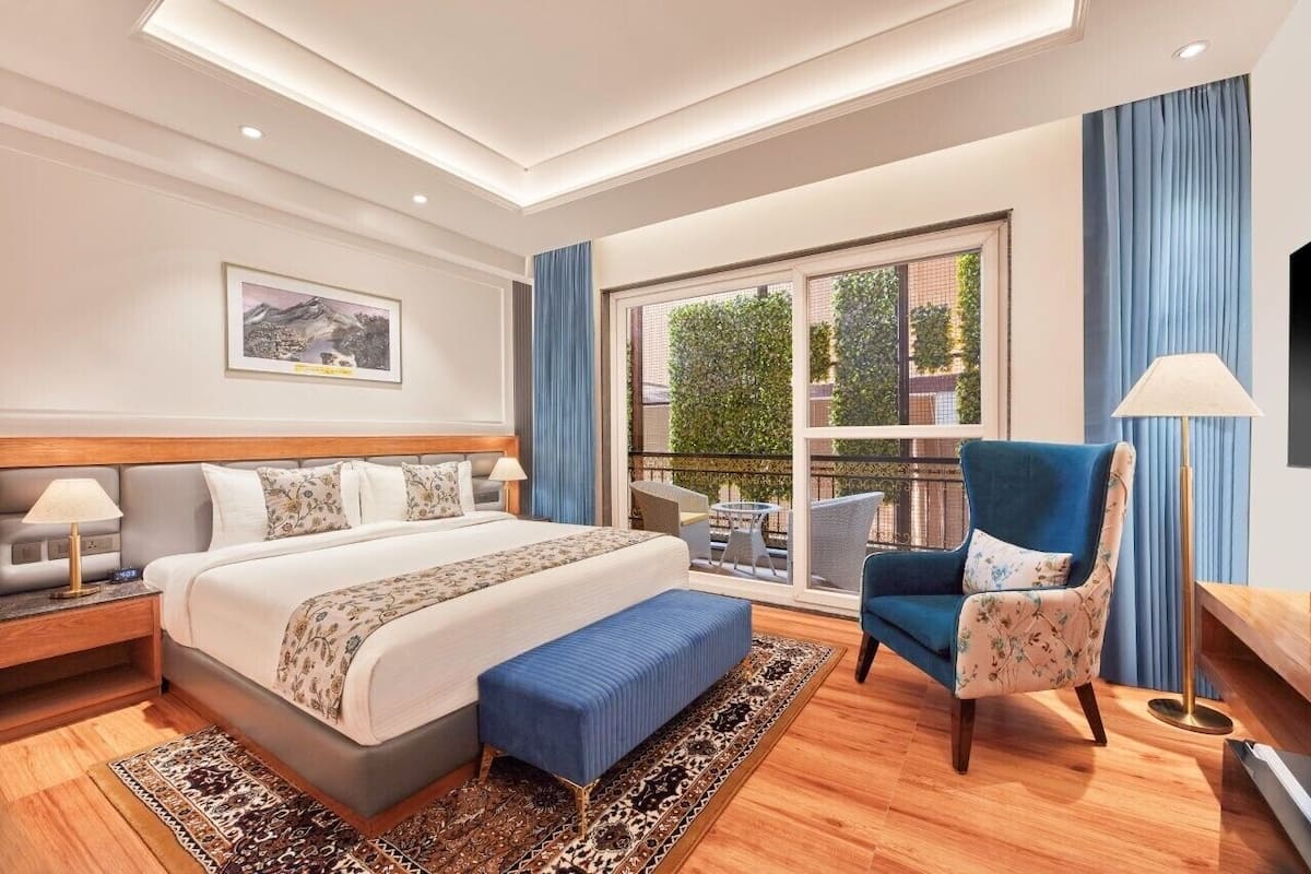 Een moderne hotelkamer met een groot bed, een zithoek met een blauwe fauteuil, een klein tafeltje en een balkon met uitzicht op groen.