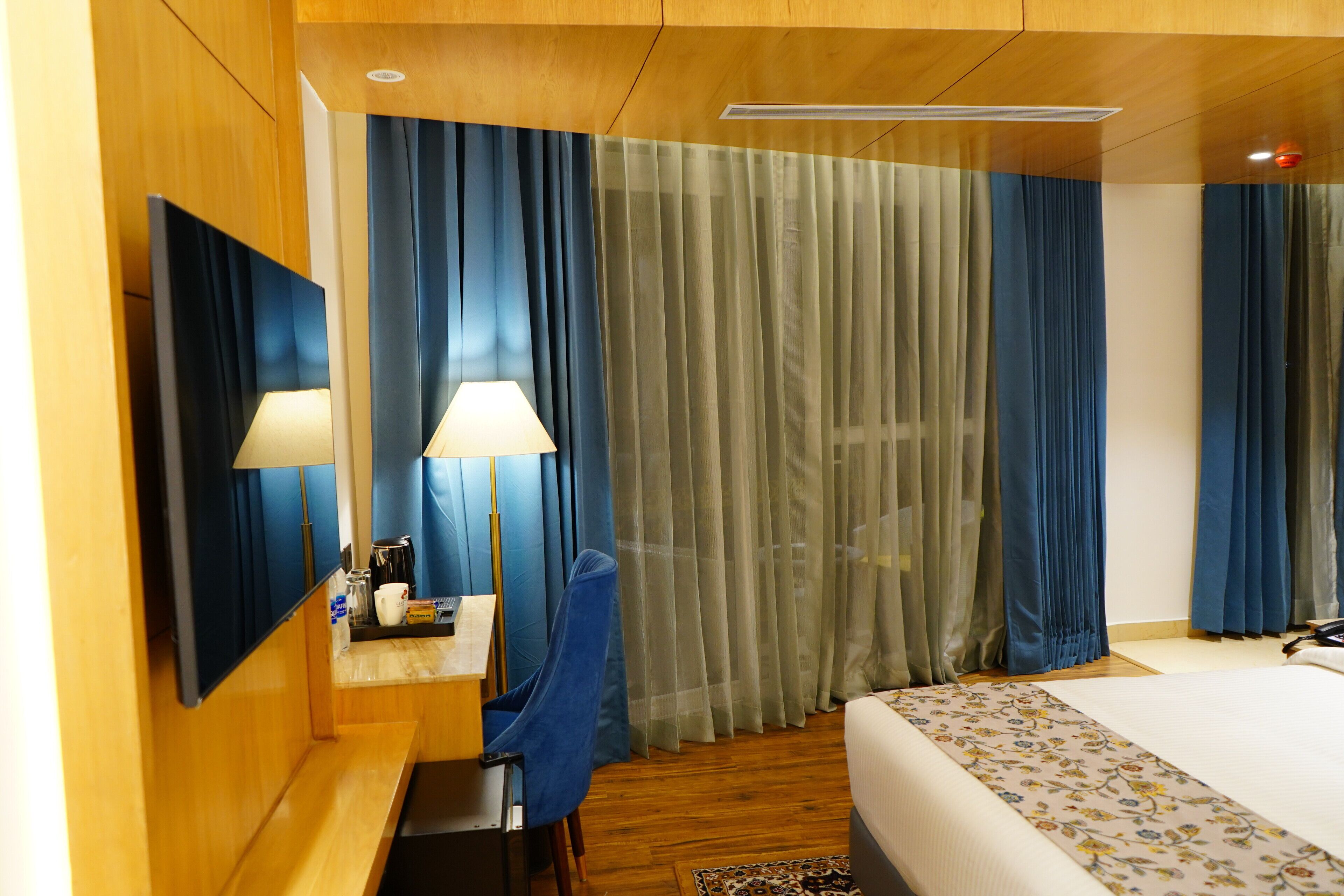 A hotel room with a bed, a desk with a chair, a mirror, and a window with curtains.