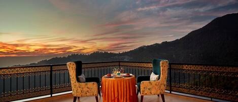 A balcony with a table set for two, overlooking a mountainous landscape during sunset.