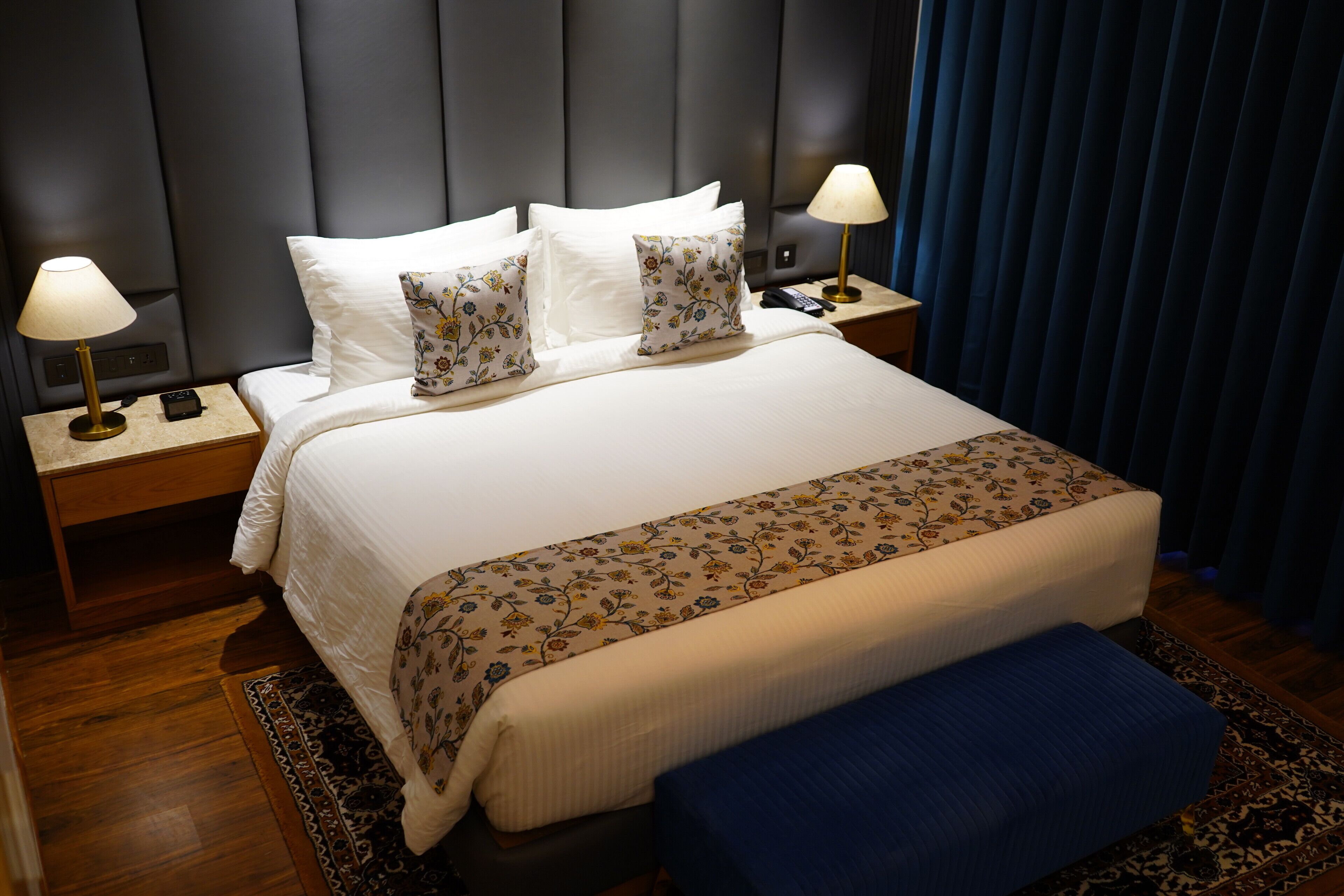 A neatly made bed with white linens and floral-patterned pillows, a blue bench, and two bedside tables with lamps.