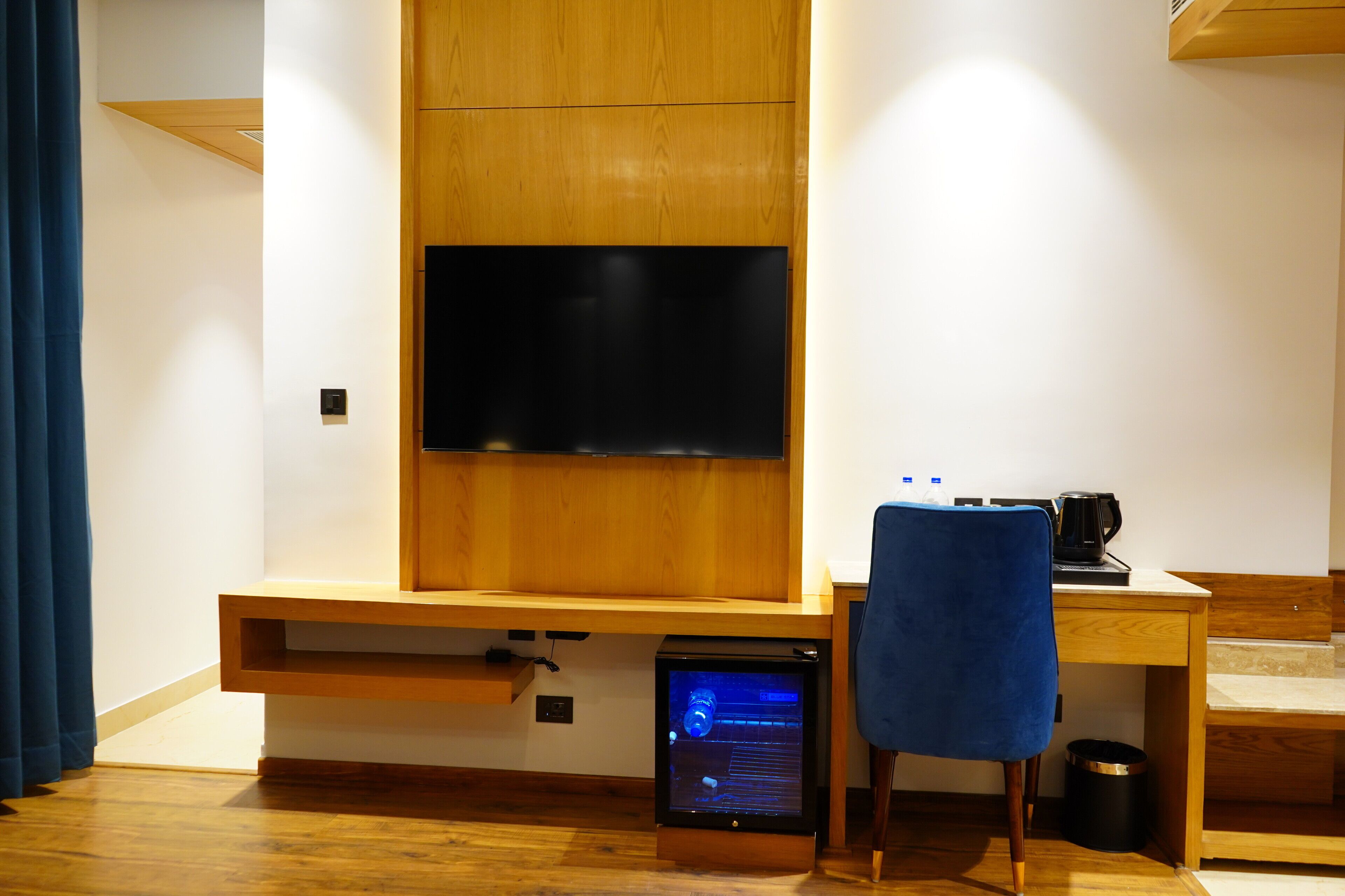 A modern room with a wooden desk, a blue chair, a television mounted on the wall, and a small computer on the desk.