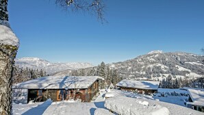 Une cabane en rondins dont le toit est recouvert de neige, avec un banc en bois, et une vue dégagée sur des montagnes enneigées en arrière-plan.