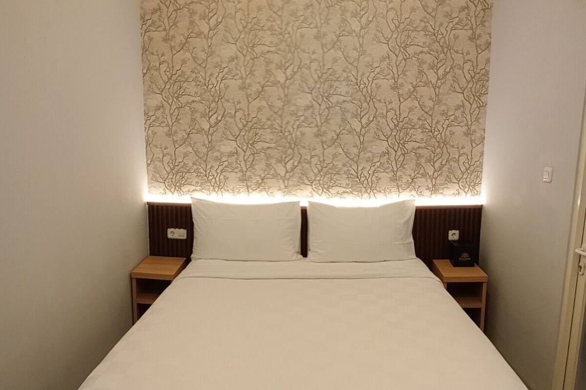 Una cama bien hecha con sábanas blancas, dos mesitas de noche con lámparas, y una pared con un diseño de árboles.