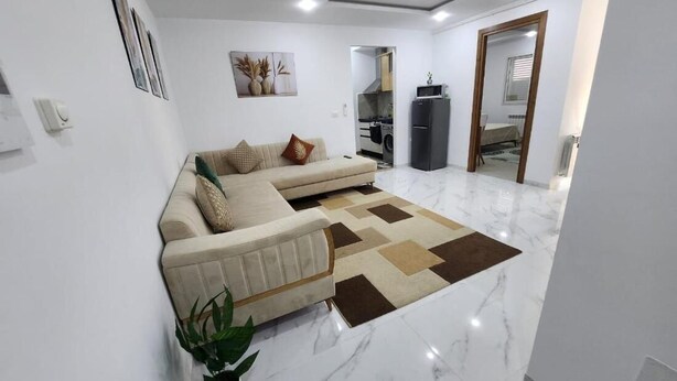 A modern living room with a sofa, a potted plant, and a kitchen in the background. - Elegant Furnished S1 With Security in Ariana - Tunisia (La Soukra)
