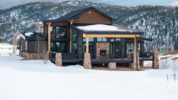 A modern two-story lodge with a stone fireplace and large windows, surrounded by snow-covered mountains. - Breathtaking Mountain Manor, Huge Deck w/ Hot Tub, Everything High End and New, 1 Mile to Ski Slopes (Granby)