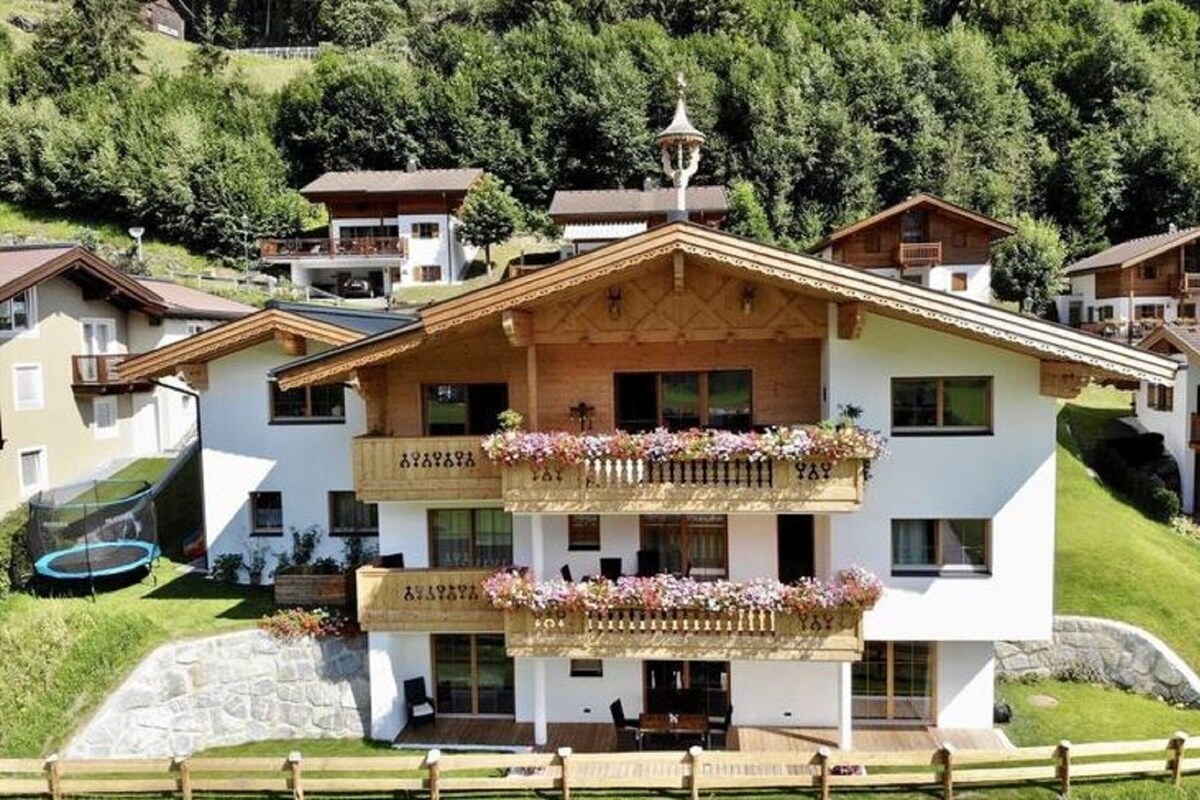 Un immeuble à plusieurs étages, doté de balcons, d’une fondation en pierre et offrant une vue sur une colline verdoyante.