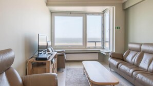 A modern living room with a large window, leather sofas, a wooden coffee table, and a TV on a stand. - Apartment With Marina View (Ostend)