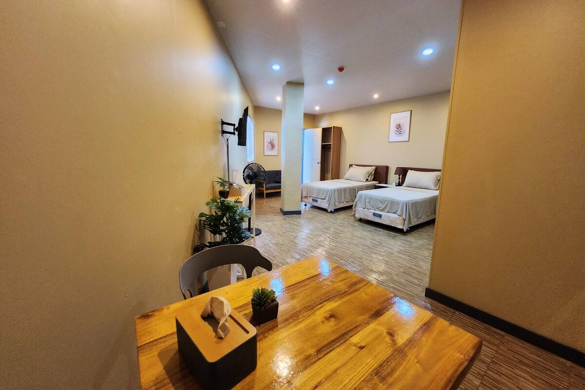 Une chambre d’hôtel avec une table en bois, deux lits, une armoire et un ventilateur.