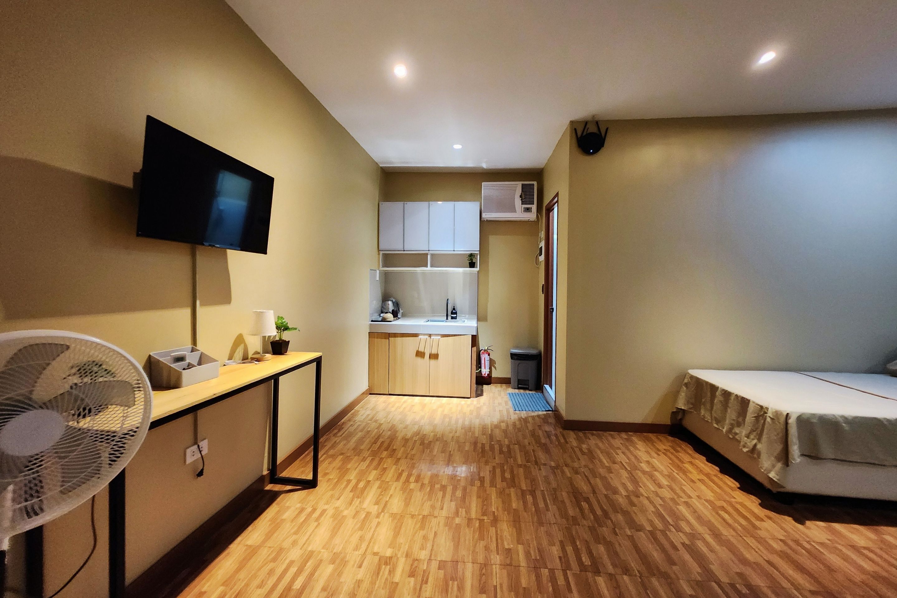 Una habitación compacta con cama, escritorio, televisor, ventilador, kitchenette y baño pequeño.