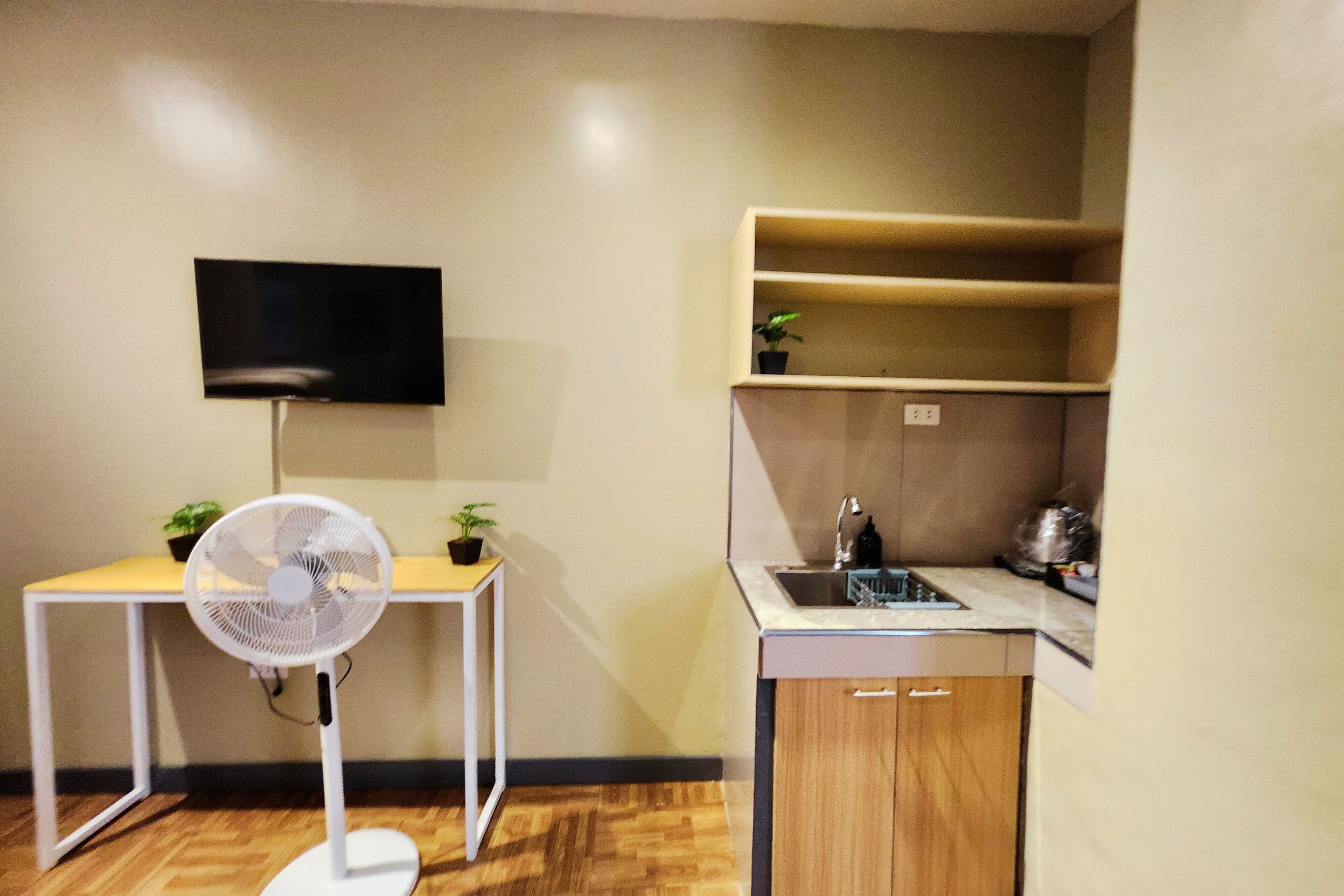 Cocina compacta con un lavamanos, una mesita con un ventilador y un televisor montado en la pared.