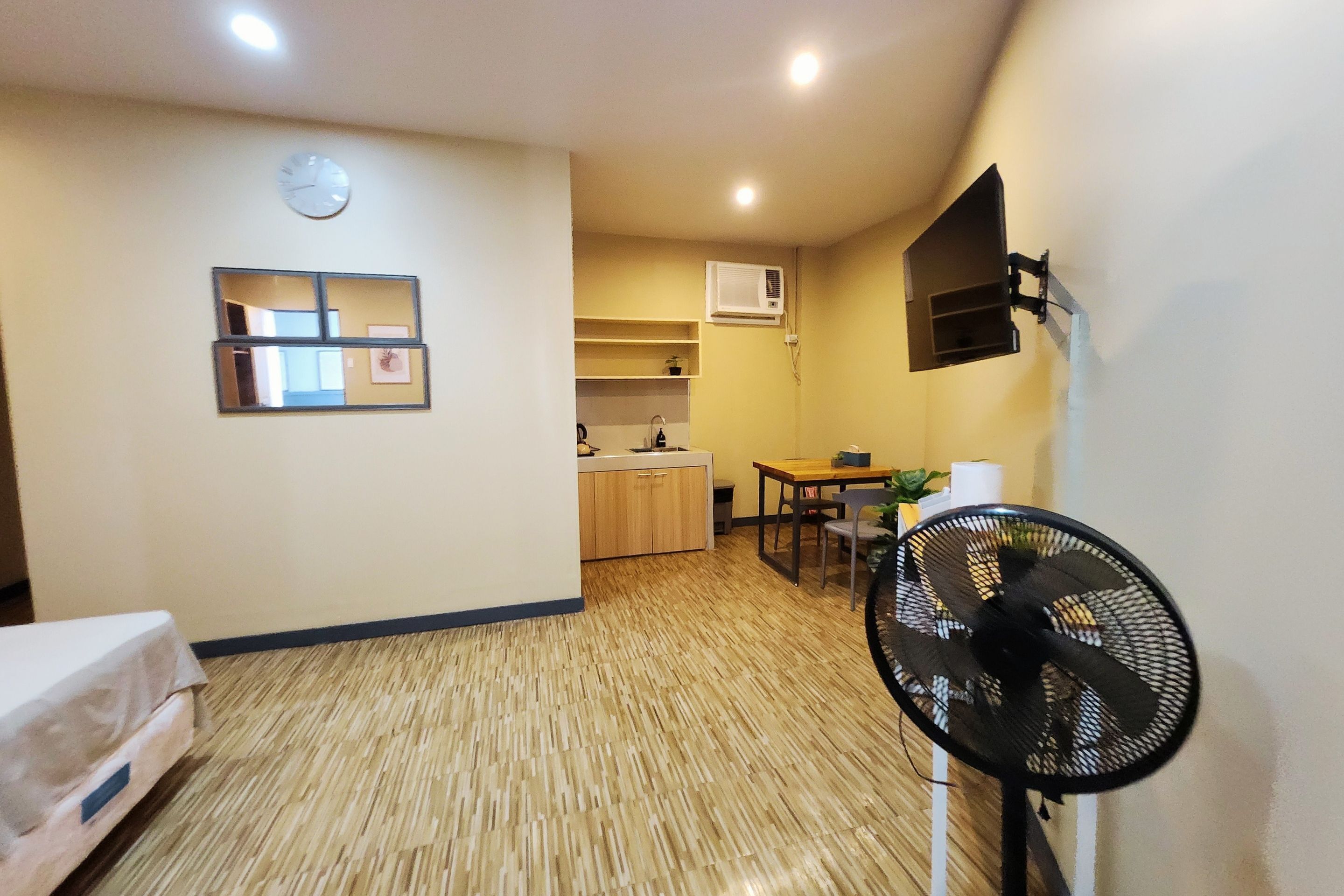 Una habitación pequeña con cama, ventilador, mesita y un rincón con kitchenette.