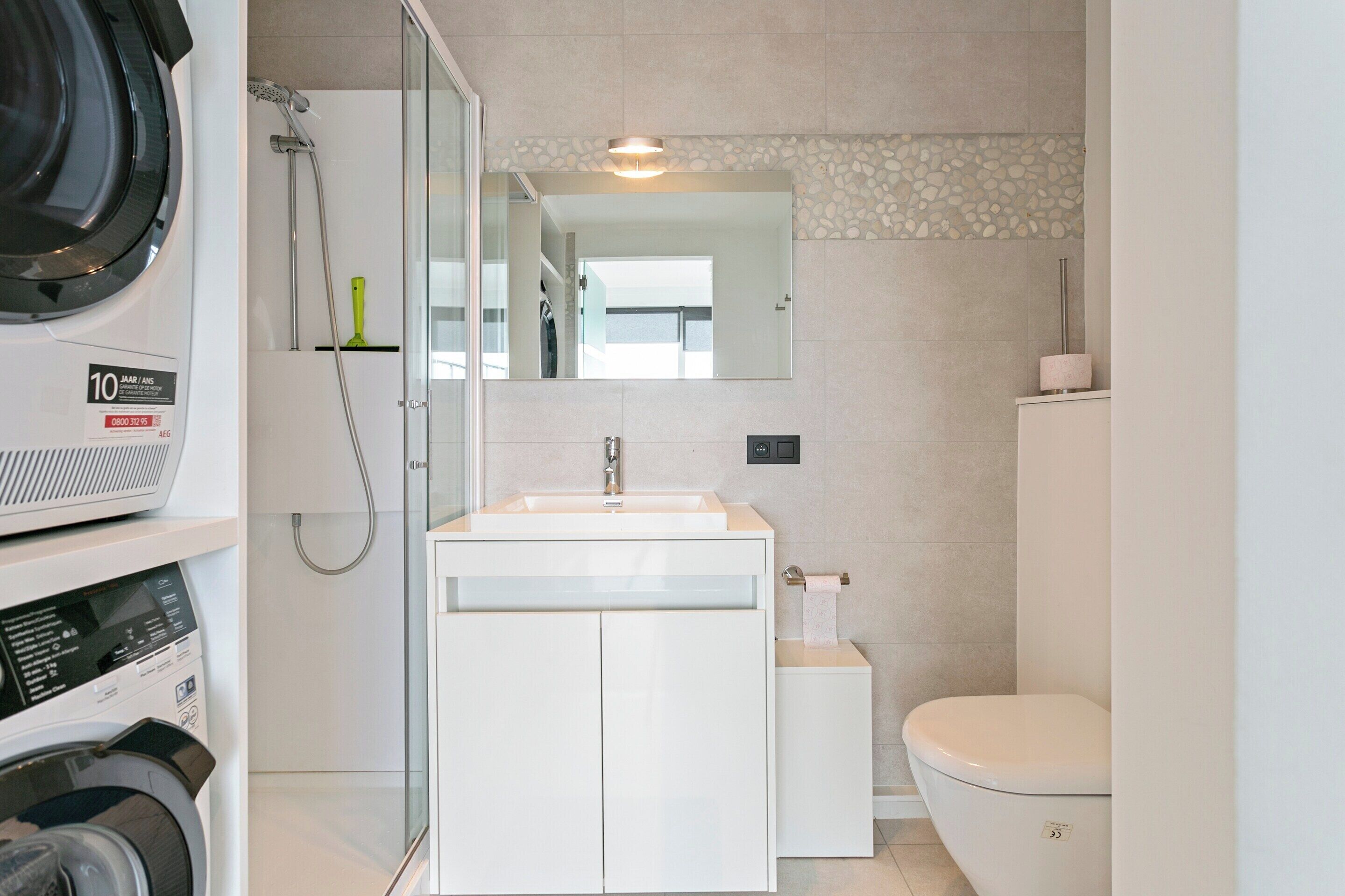 Un baño moderno con lavadora integrada, ducha con mampara de vidrio, lavabo blanco y retrete.