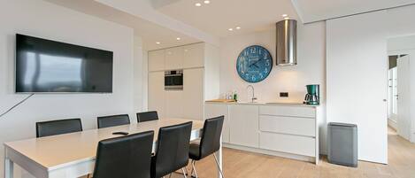 A modern kitchen with a dining area, white cabinetry, a large clock, and a flat-screen TV.