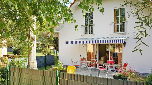 Ein zweistöckiges Haus mit einer Terrasse, die einen Tisch, Stühle, eine überdachte Sitzgruppe und einen umzäunten Garten umfasst.