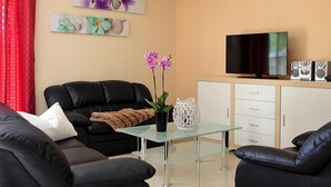 Ein Wohnzimmer mit Glastisch, schwarzer Ledersofa, Flachbildfernseher und einer Anrichte mit Lautsprechern.