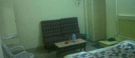 A small, sparsely furnished room with a plaid sofa, a small table with a water bottle, a bed with a patterned bedspread, and a white chair.